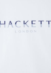 Bílá látka s nápisem "HACKETT" v tučných přechodech modré barvy a "LONDON" v menším písmu níže. Hladká textura s moderním designem.