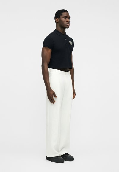 Un homme debout, tourné vers la droite, porte un polo noir raccourci, un pantalon blanc à jambes larges et des baskets noires sur un fond uni.