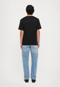 Hombre con cabello negro rizado que lleva una camiseta negra lisa, jeans de mezclilla claros y zapatos negros, de pie de espaldas sobre un fondo blanco.