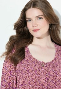 Blomsterbluse i lyserøde og lilla nuancer, der har et struktureret stof, rund halsudskæring og en frontbinding. Lange ærmer.