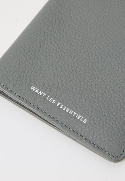 Portefeuille en cuir gris avec une finition texturée. Il présente un texte blanc en relief "WANT LES ESSENTIELS" en bas. Bords soigneusement cousus.