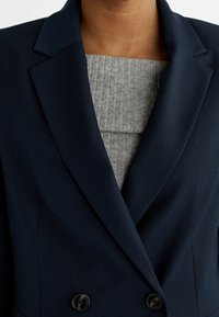 Blazer doppiopetto blu navy realizzato in tessuto liscio con revers strutturati, dotato di due bottoni neri e un top grigio a coste sottostante.