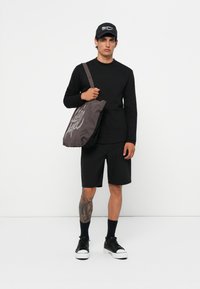 Svart langermet skjorte, svarte shorts, og svarte joggesko med hvite såler. Grå totebag med stilisert tekst, båret på skulderen.