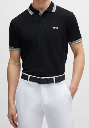 Schwarzes Poloshirt mit einem Kragen mit grauen und weißen Streifen, zwei Knöpfen und dem 'BOSS'-Logo. Kombiniert mit einem schwarzen Gürtel und weißen Hosen.