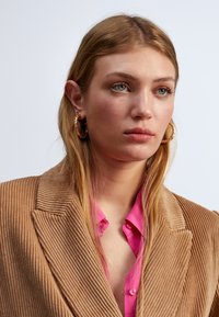 Il modello indossa un blazer in corduroy rigato color beige e una camicia button-up rosa brillante, abbinati a grandi orecchini a cerchio d'oro.