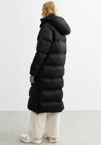 Abrigo de plumas negro con capucha, con un diseño largo y acolchado y cremalleras laterales, acompañado de pantalones anchos blancos y zapatillas chunky.