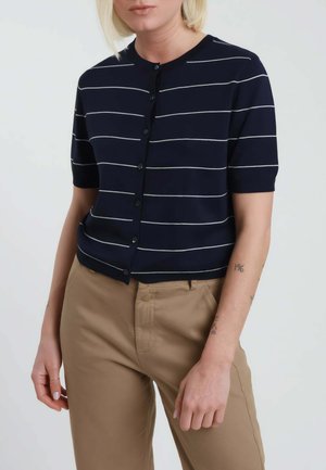 Donna che indossa una maglia blu navy a maniche corte con bottoni e sottili strisce orizzontali bianche, e pantaloni marrone chiaro su uno sfondo neutro.