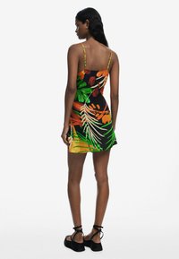 Vestido negro con patrones de hojas tropicales coloridas, tirantes finos y dobladillo acampanado, combinado con sandalias de plataforma negras. Textura de tela suave.