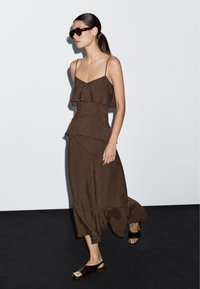 Hněd dresses s vrstvami a tenkými ramínky, splývavá látka a volánkový design. V páru s tmavými sandály s otevřenou špičkou a slunečními brýlemi.