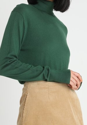 Personne portant un pull à col roulé vert et une jupe en velours côtelé beige, les mains délicatement jointes près de la taille.