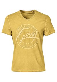 Gul kortærmet V-hals T-shirt med et falmet cirkulært logo "Guess Los Angeles California est. 1981" trykt på forsiden.