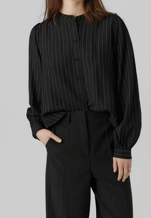 Femme portant un chemisier noir à manches longues boutonné avec des fines rayures blanches et un pantalon noir taille haute, une main dans la poche.