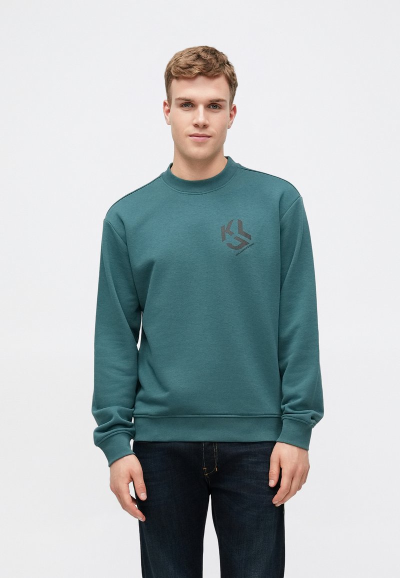 Teal sweatshirt laget av bomull-blandingsstoff, har rund hals og ribbede ermer, med en svart logo på venstre bryst. Kombinert med mørke jeans.