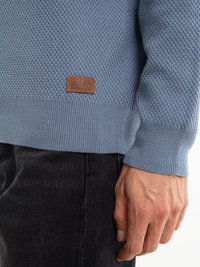 Gai zils teksturēts džemperis ar ribotām piedurknēm un apakšmalu. Iekļauts brūns ādas logotipa uzlīmē, kas saskaņots ar tumšiem teksturētiem biksēm.