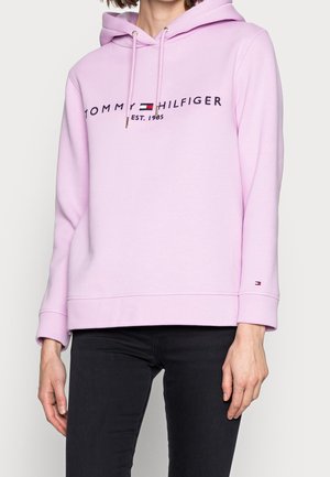 Kobieta w jasnoróżowej bluzie z kapturem Tommy Hilfiger z logo i napisem na piersi, w czarnych spodniach, stojąca na jednolitym tle.