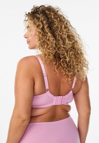 Mujer con cabello rizado vista desde atrás, vistiendo un sujetador rosa claro y ropa interior a juego sobre un fondo blanco.