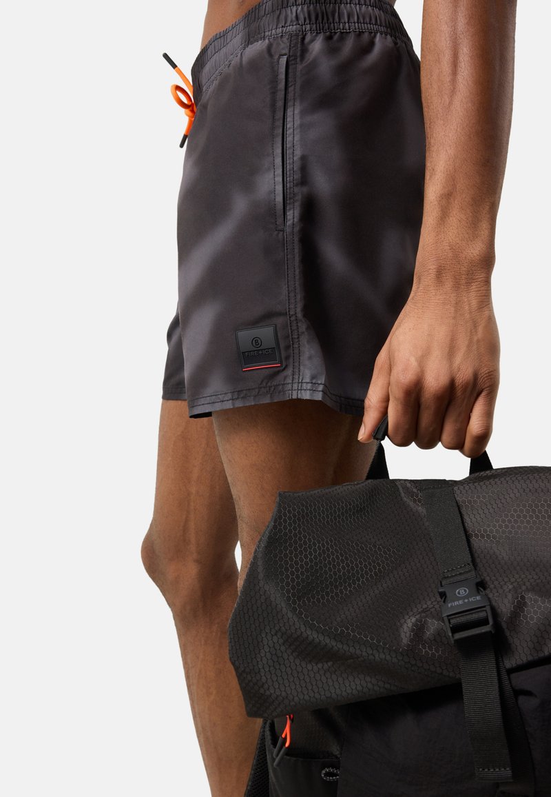 Homme portant un short de bain gris foncé avec un cordon orange, tenant un sac à dos noir texturé par sa poignée supérieure.