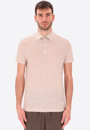 Giovane uomo con capelli corti castani che indossa una polo beige a maniche corte e pantaloni marroni, in piedi davanti a uno sfondo chiaro e semplice.
