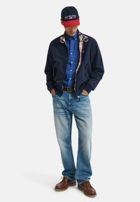 Giacca blu navy con fodera a quadri, camicia blu, jeans azzurri e scarpe marroni. Cappellino da baseball presenta colori rosso e blu navy con logo.