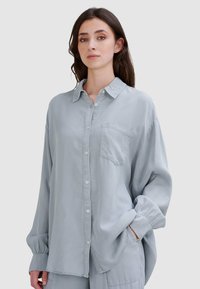 Jasnoniebieska koszula z długim rękawem z guzikiem z przodu i kieszenią na klatce piersiowej. Posiada luźny krój i miękką, gładką teksturę.