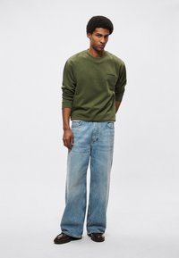 Sudadera verde oliva con cuello redondo y puños acanalados, combinada con vaqueros de pierna ancha de color azul claro. La modelo está de pie frente a un fondo liso.