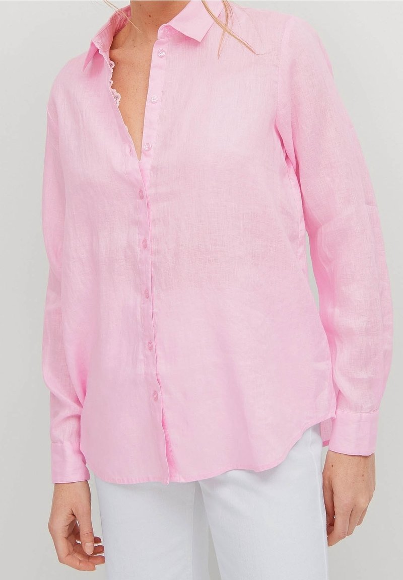 Femme portant une chemise rose pâle à manches longues boutonnée et un pantalon blanc, debout devant un fond clair uni.