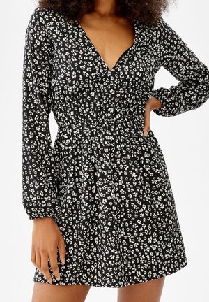 Schwarzes Kleid mit kleinem weißem Blumenmuster, langen Ärmeln, elastischer Taille und V-Ausschnitt, getragen von einer Person mit der Hand in der Hüfte.