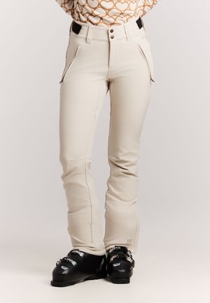 Person iført beige skibukser og sorte skistøvler står mod en neutral baggrund.