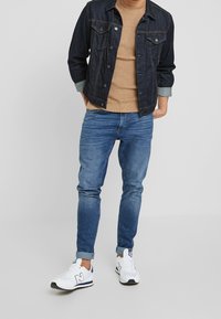 Dark denim jacket over a beige knit sweater, blue slim-fit jeans, and white athletic shoes with navy accents. Casual streetwear look.