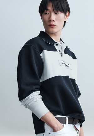 Νέος άνδρας με μαύρα μαλλιά που φοράει μακρυμάνικο ράγκμπι μπλουζάκι ασπρόμαυρο, κοιτάζει προς τα πλάγια με το ένα χέρι στην τσέπη λευκού παντελονιού.