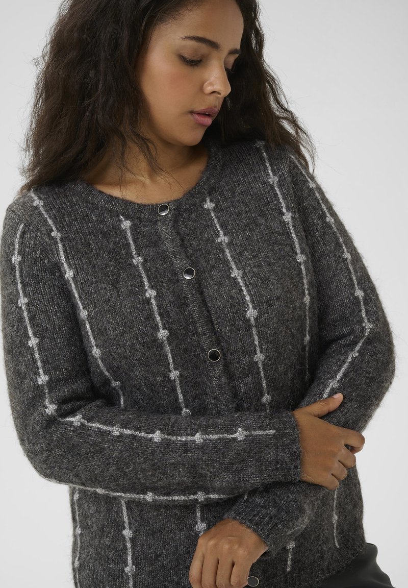 Femme aux longs cheveux bruns portant un gilet en tricot gris foncé avec des rayures blanches verticales et des boutons noirs ronds, regardant vers le bas et tenant son bras.