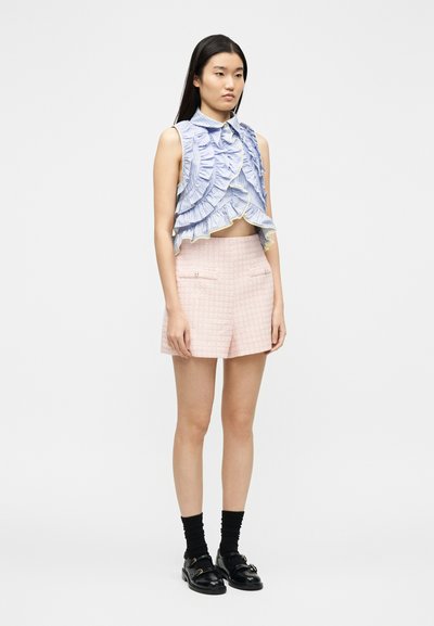 Νεαρή γυναίκα που φοράει μια αμάνικη μπλούζα με βολάν σε μπλε χρώμα, ροζ σορτς με υφή, μαύρες κάλτσες και μαύρα μοκασίνια, στέκεται μπροστά σε απλό λευκό φόντο.