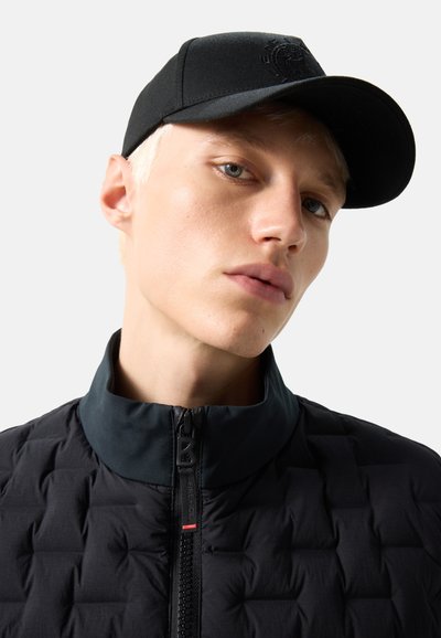 Junge Person mit kurzen blonden Haaren, die eine schwarze Baseballkappe und eine gesteppte schwarze Jacke mit Reißverschlusskragen trägt.