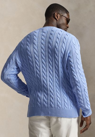 Muž v svetlomodrom vrkočišom svetri a bielych nohaviciach, otočený chrbtom, s jednou rukou v bok na neutrálom pozadí.