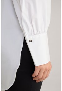 Camicia bianca a maniche lunghe con dettagli a polso arricciati e accento di bottone metallico. Tessuto morbido, design svasato e spacchi laterali per maggiore facilita.
