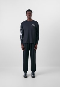 Schwarzes Langarmshirt mit grafischen Designs auf den Ärmeln und Logo auf der Brust. Kombiniert mit locker sitzenden schwarzen Hosen und Sportschuhen.