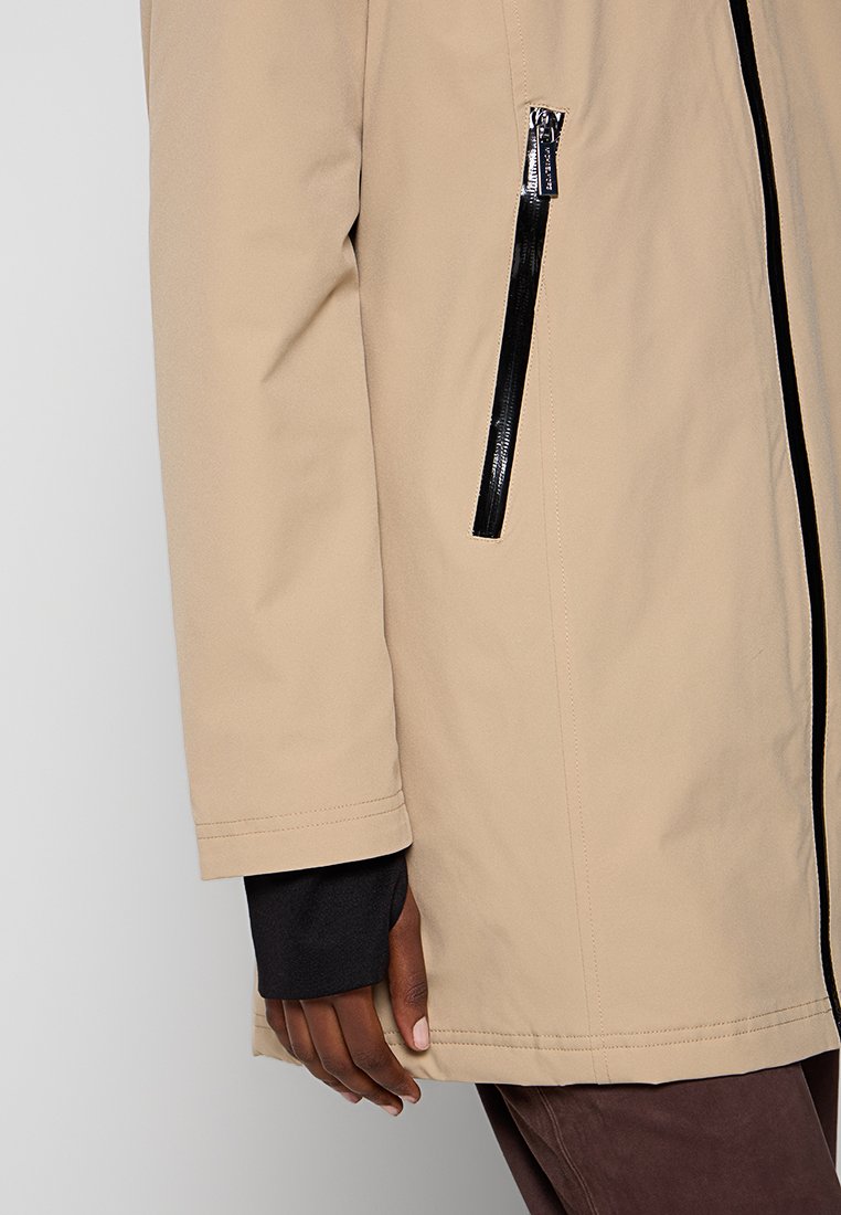 Beige jas met een gladde textuur, voorzien van een zwart ritszakje aan de linkerkant en een contrasterende zwarte binnenlaag.