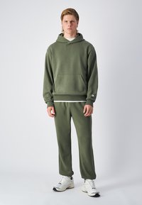 Sudadera verde oliva con un bolsillo frontal y puños acanalados, combinada con pantalones de chándal a juego. El modelo lleva zapatillas blancas.