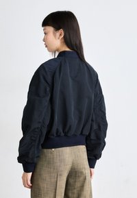 Marineblauwe bomberjack met opstaande mouwen, ribbelmanchetten en een aansluitende tailleband. Het jack heeft een gladde textuur en een minimalistisch ontwerp.