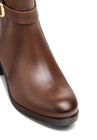 Botín de cuero marrón hasta el tobillo con punta redonda, correa decorativa, hebilla dorada y tacón bajo negro sobre un fondo blanco.