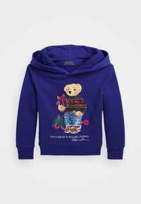 Felpa con cappuccio viola con grafica di un orso che indossa un maglione nero e jeans blu, che tiene un regalo. L'illustrazione include elementi festivi.
