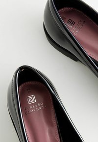 Scarpe in vernice nera con punta arrotondata e soletta imbottita bordeaux. La soletta presenta il marchio "Forever Comfort".