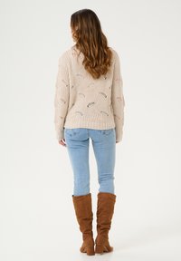 Pull en tricot beige avec un motif ajouré, associé à un jean skinny bleu clair et des bottes marron hauteur genou. Design simple et texturé.