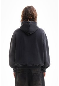 Personne aux cheveux bruns ondulés mi-longs, portant un sweat à capuche noir et un pantalon noir, debout de dos sur un fond blanc.