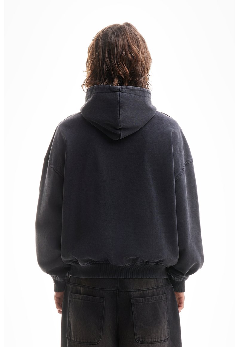 Personne aux cheveux bruns ondulés mi-longs, portant un sweat à capuche noir et un pantalon noir, debout de dos sur un fond blanc.