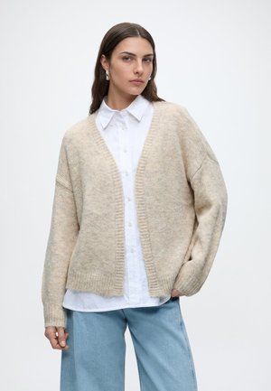 Femme aux longs cheveux bruns portant des boucles d'oreilles argentées en forme d'anneaux, un cardigan beige par-dessus une chemise blanche boutonnée et un jean bleu clair, debout sur un fond blanc.