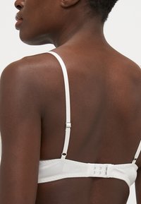 Soutien-gorge blanc avec bretelles ajustables, texture de tissu lisse, fermeture à agrafes et détails de design subtils sur la bande. Vue arrière affichée.