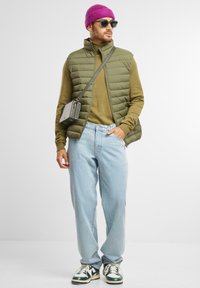 Olivegrüste Steppweste über einem braunen Rollkragenpullover, hellblaue Jeans, schwarze Umhängetasche, lila Strickmütze und schwarz-weiße Sneakers.