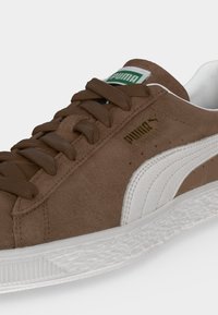 Καφέ σουέτ αθλητικό παπούτσι Puma με λευκή πλευρική ρίγα και υφή στη λευκή σόλα, που δείχνει τρύπες για κορδόνια, κορδόνια και λογότυπο της μάρκας στο πλάι και στη γλώσσα.
