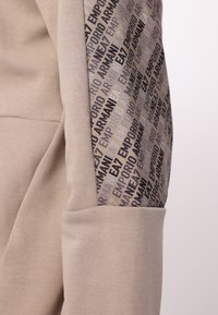 Sudadera color beige con una textura suave, que presenta un panel lateral con un diseño en gris y "Emporio Armani" en varios tonos.